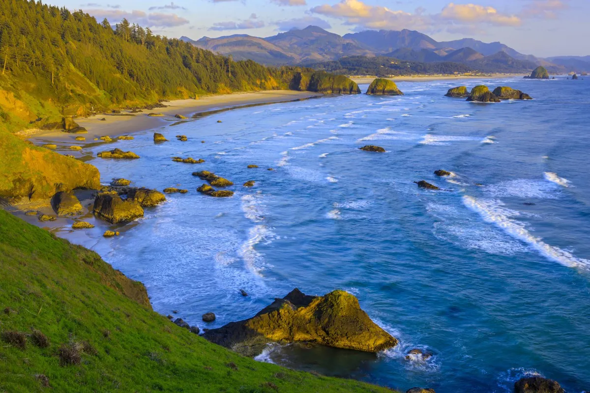 A panoramic view of a rocky beach framed by gentle waves and distant mountains, with lush greenery bordering the scene. Visitors are depicted beachcombing, appreciating the sunset, and exploring the serene coastline.
