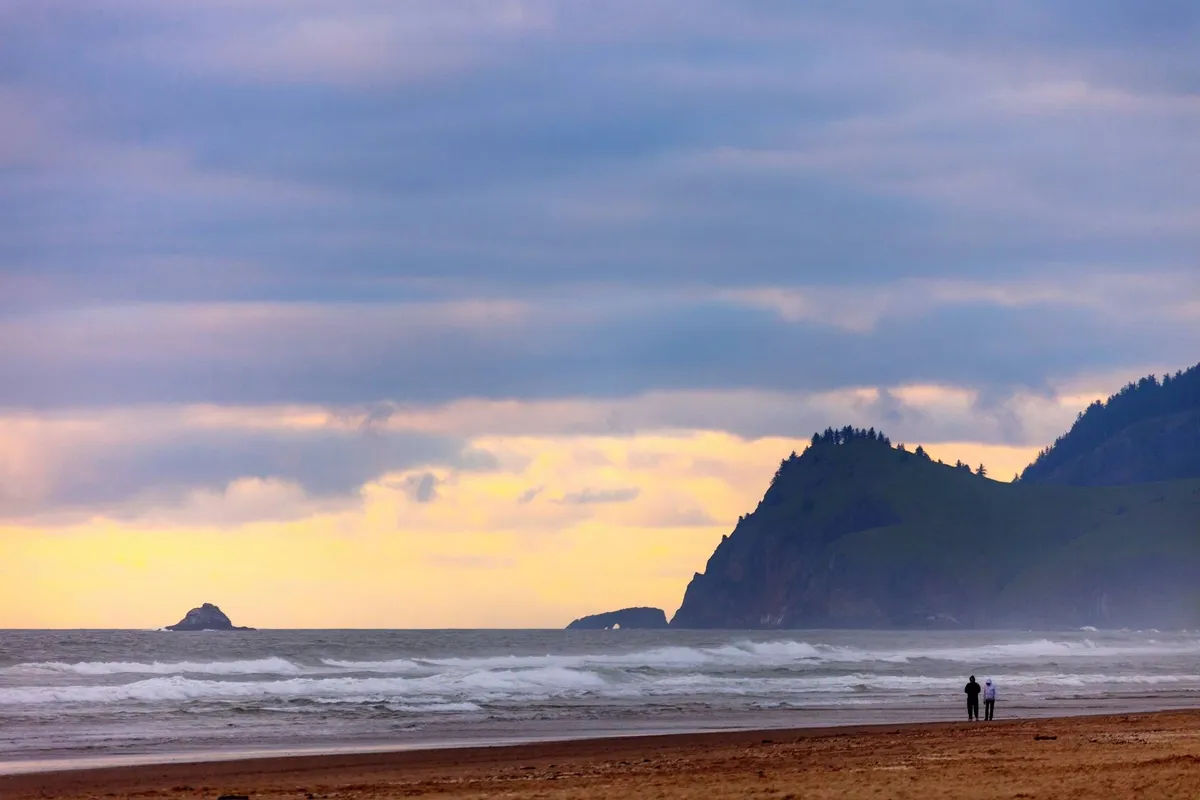 A tranquil beach scene features gentle ocean waves lapping against the sand, distant cliffs forming a dramatic backdrop, and a couple strolling hand-in-hand near a distinctive rock formation. The mood is peaceful and romantic, with the promise of beachcombing and sunset walks.