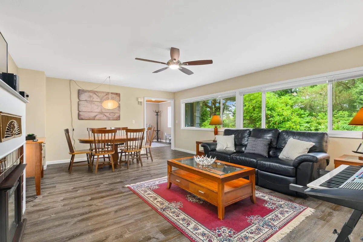 A spacious living area with a comfortable sofa, dining table, large windows letting in abundant natural light, and a coffee table. The room is arranged for both relaxation and socializing, with views of the lush greenery outside through the panoramic windows. The inviting atmosphere encourages gathering for movies, music, or a cozy evening in.