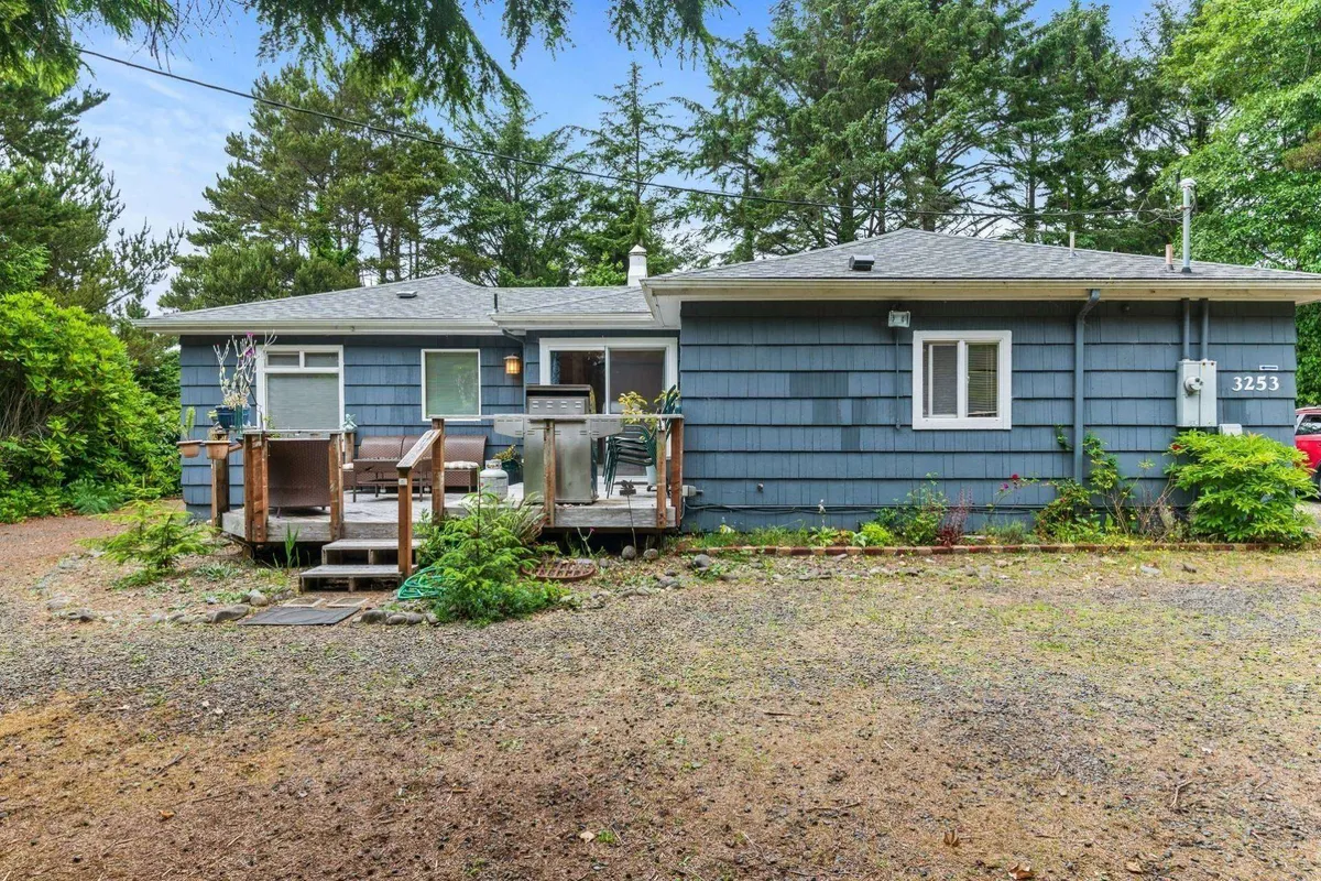 A tranquil wooden deck bordered by lush greenery, with blue exterior walls visible. The space is set up for relaxation or gatherings—ideal for a family BBQ after time at the beach. The outdoor deck suggests a private retreat for meals and socializing.