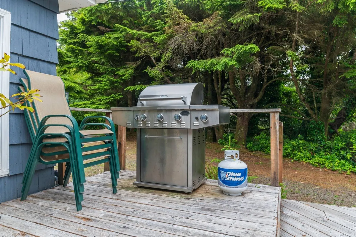 An inviting exterior deck is set amidst lush greenery, with a grill and a stack of chairs hinting at leisurely barbecues and outdoor gatherings. The view emphasizes relaxation and the social aspect of spending time outdoors, surrounded by nature.