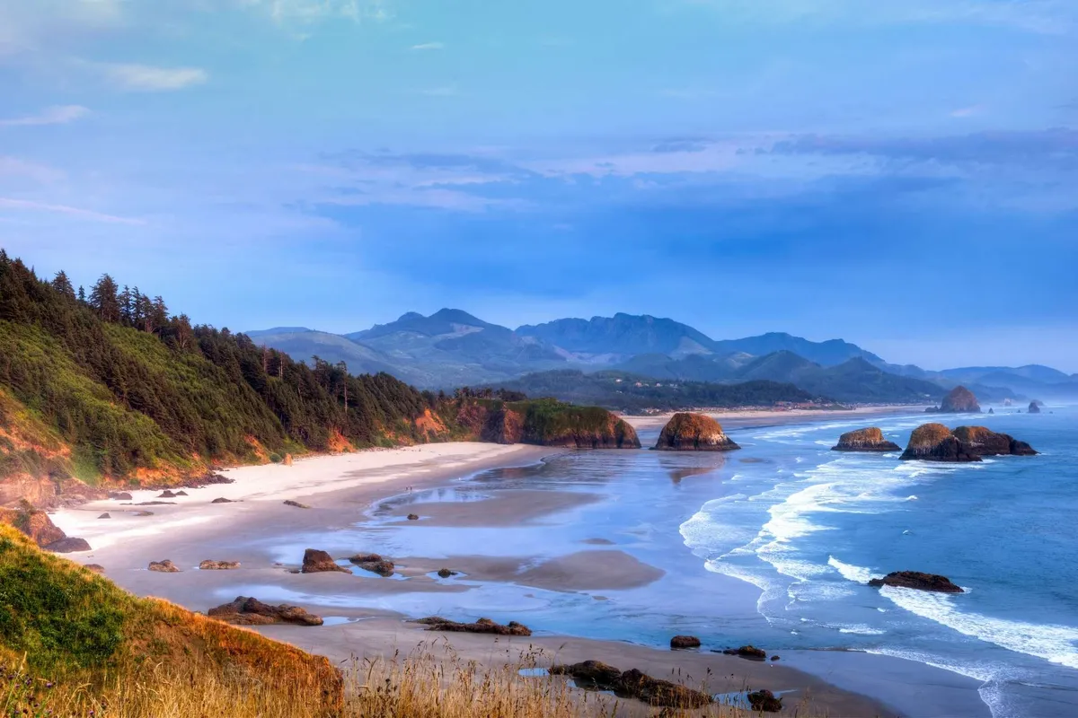 A sweeping view of the beach with sandy shores, striking rock formations, the ocean, distant mountains, and an expansive sky. The image captures a tranquil mood, ideal for beach outings, walking, and exploration.