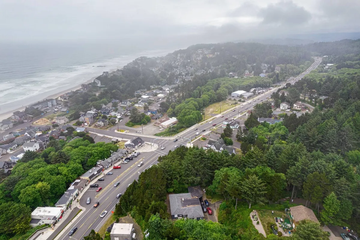 A sweeping aerial view captures a picturesque stretch of Oregon's coastline, with a winding coastal road flanked by clusters of residential homes, lush green trees, and the expansive ocean shimmering in the background. The scene feels serene, inviting viewers to imagine exploring the area, taking coastal drives, and soaking in the natural beauty.