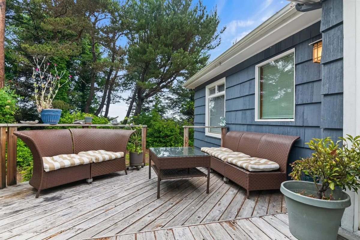 The image showcases an inviting deck furnished with woven wicker seating and a glass-top table, surrounded by lush greenery and dappled with soft sunlight filtering through the trees. The setting suggests a tranquil outdoor space perfect for relaxing, sipping coffee, or gathering with loved ones, with the promise of sunset and starlit views.