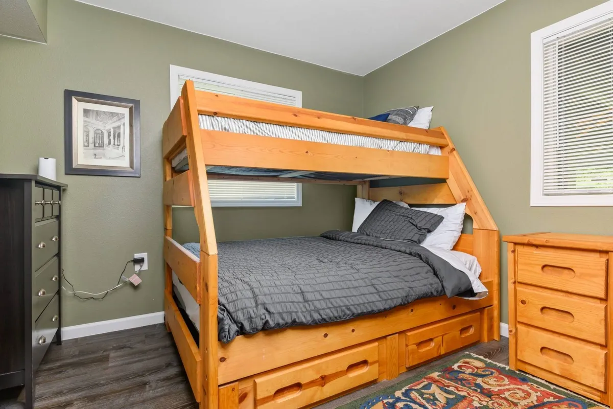 A warmly lit bedroom features a full-over-twin bunk bed, a chest of drawers, and sunlight streaming in through the window. The room looks comfortable and functional, ideal for children or siblings to share.