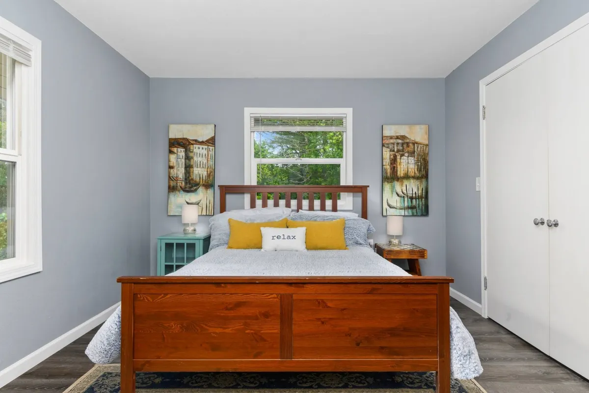 A serene bedroom centered on a wooden queen bed with 'relax' pillows, ample natural light, and tranquil wall art. The inviting setup is ideal for restful nights and peaceful mornings, suitable for adults or couples.